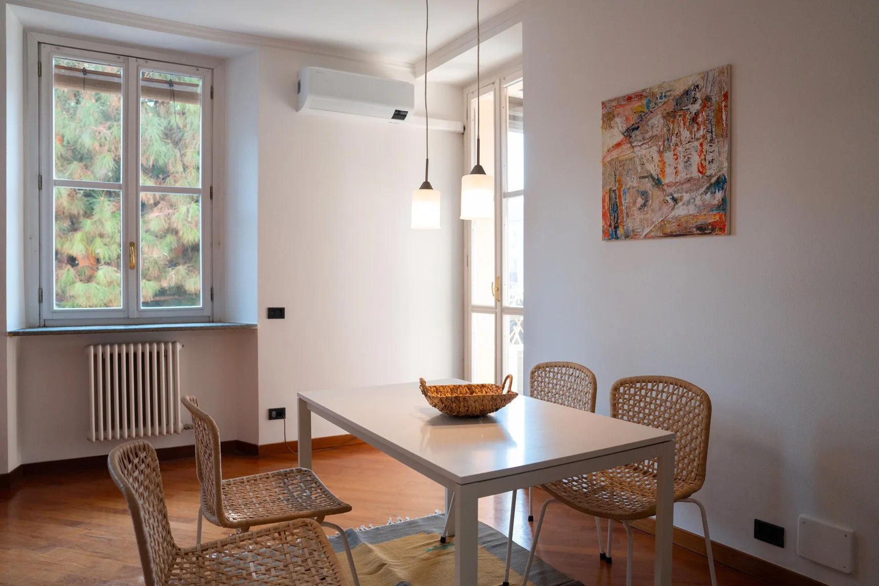 Comedor con una mesa y asientos tejidos junto a ventanas altas.