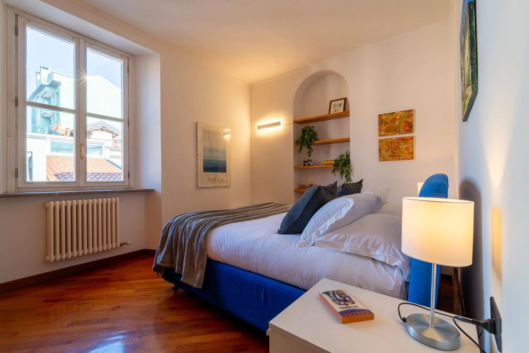 Dormitorio con una cama junto a una gran ventana y una lámpara de mesa encendida.