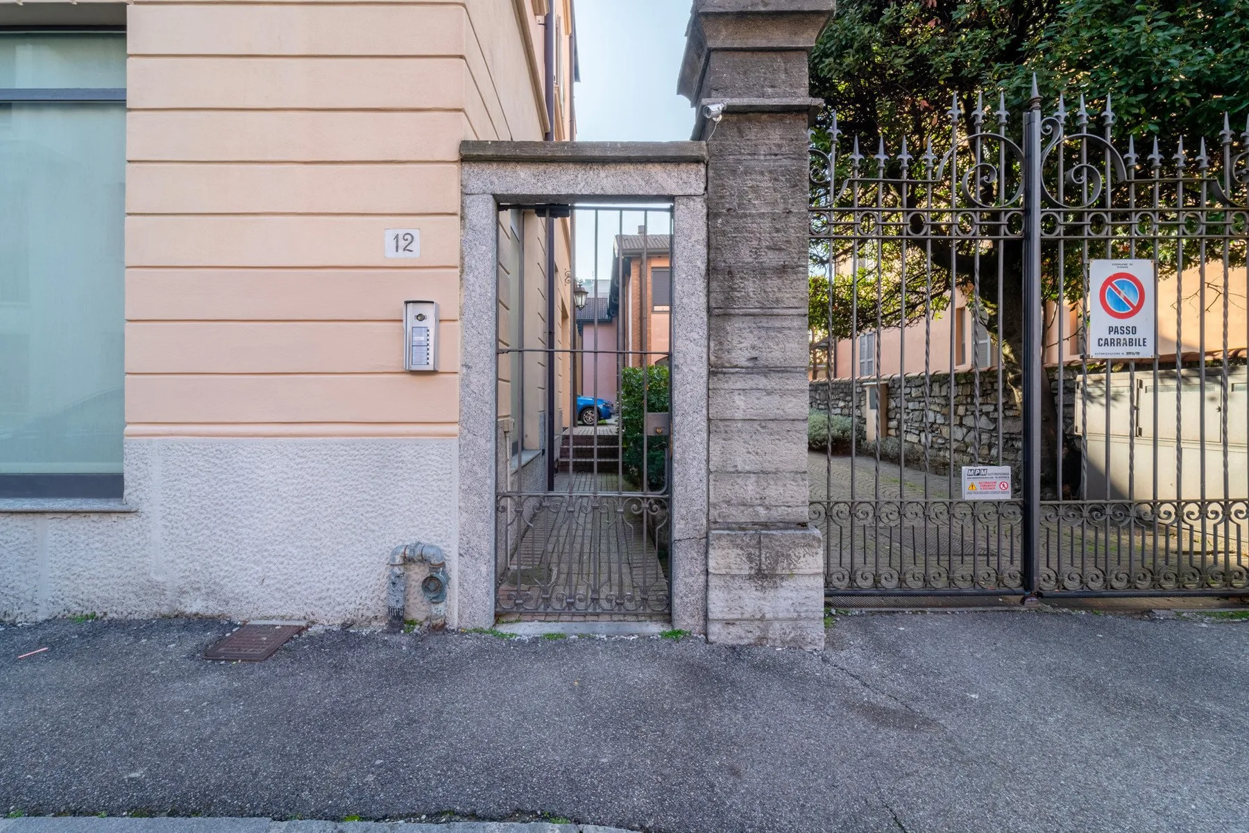 Una entrada con portón junto a un edificio claro, con una pequeña puerta peatonal y suelo pavimentado.