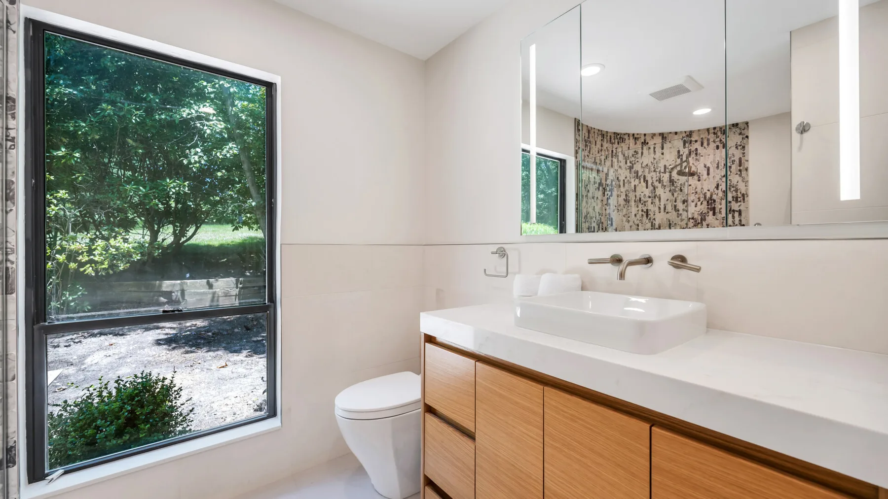 A bright bathroom has a wide vanity beside a large window overlooking greenery.
