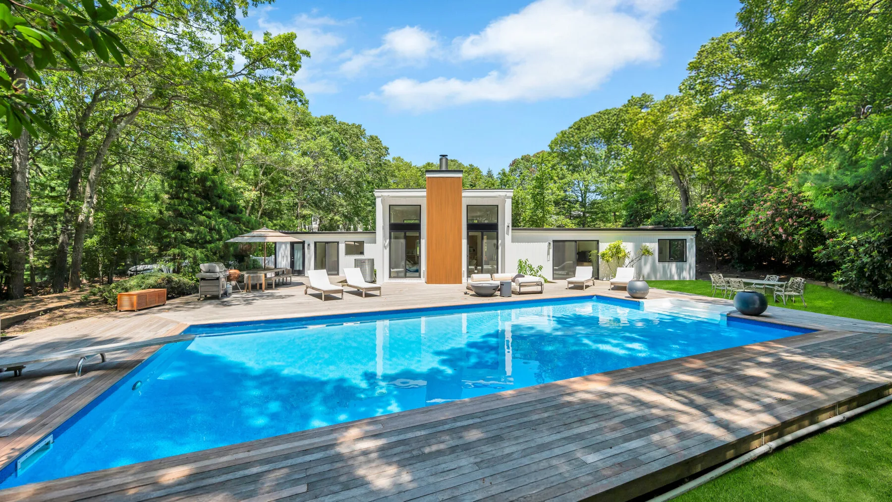 A modern house stands behind a swimming pool, surrounded by leafy trees.