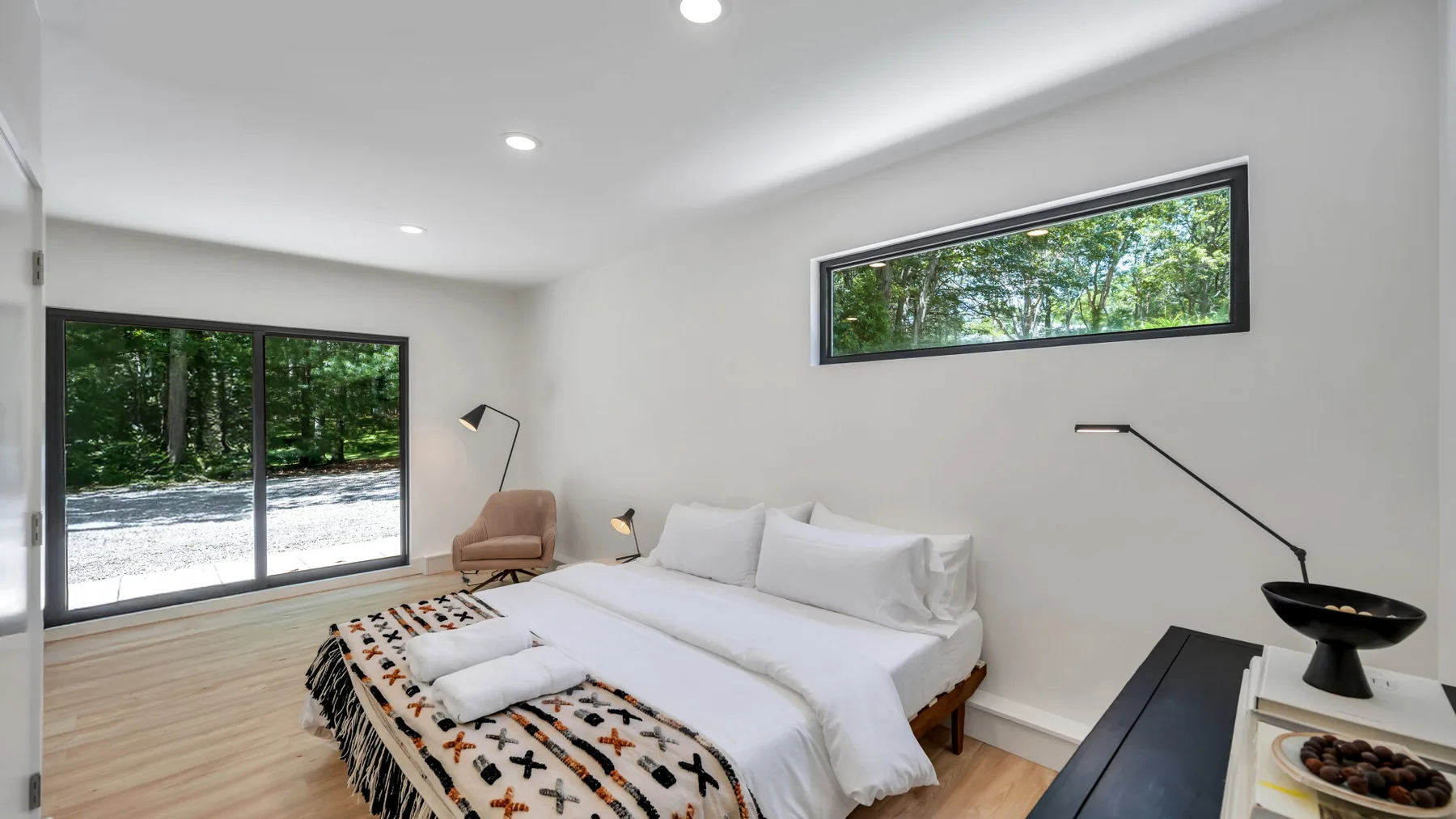 A bright bedroom with a white bed and large windows overlooking trees.
