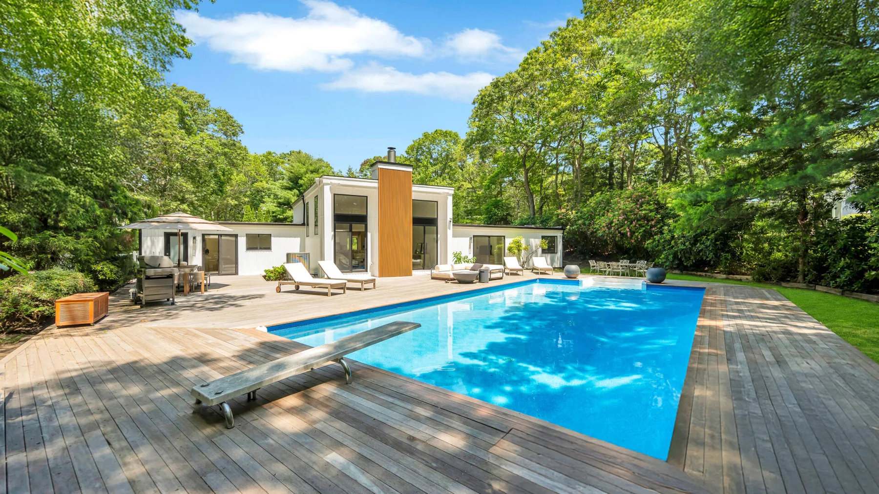 A modern house opens onto a swimming pool and a broad wooden deck amid trees.
