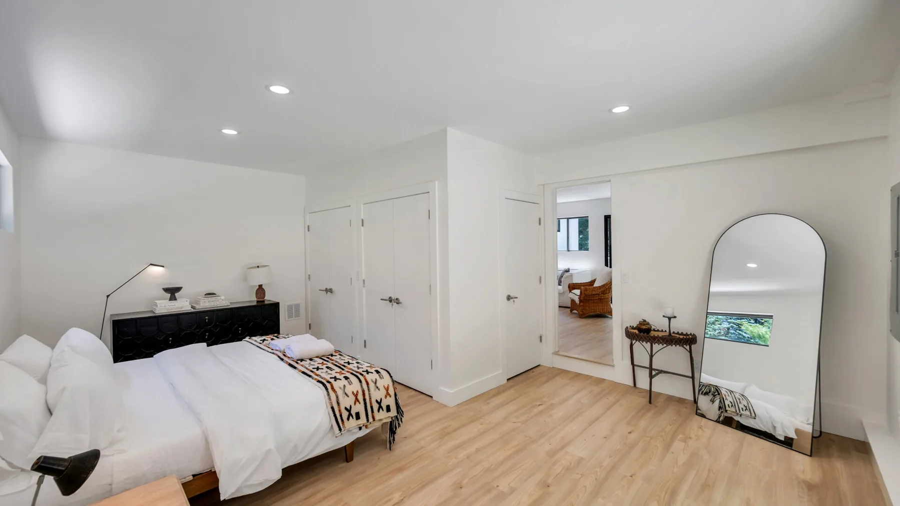 A bright bedroom with white bedding, light wood flooring, and a full-length mirror.