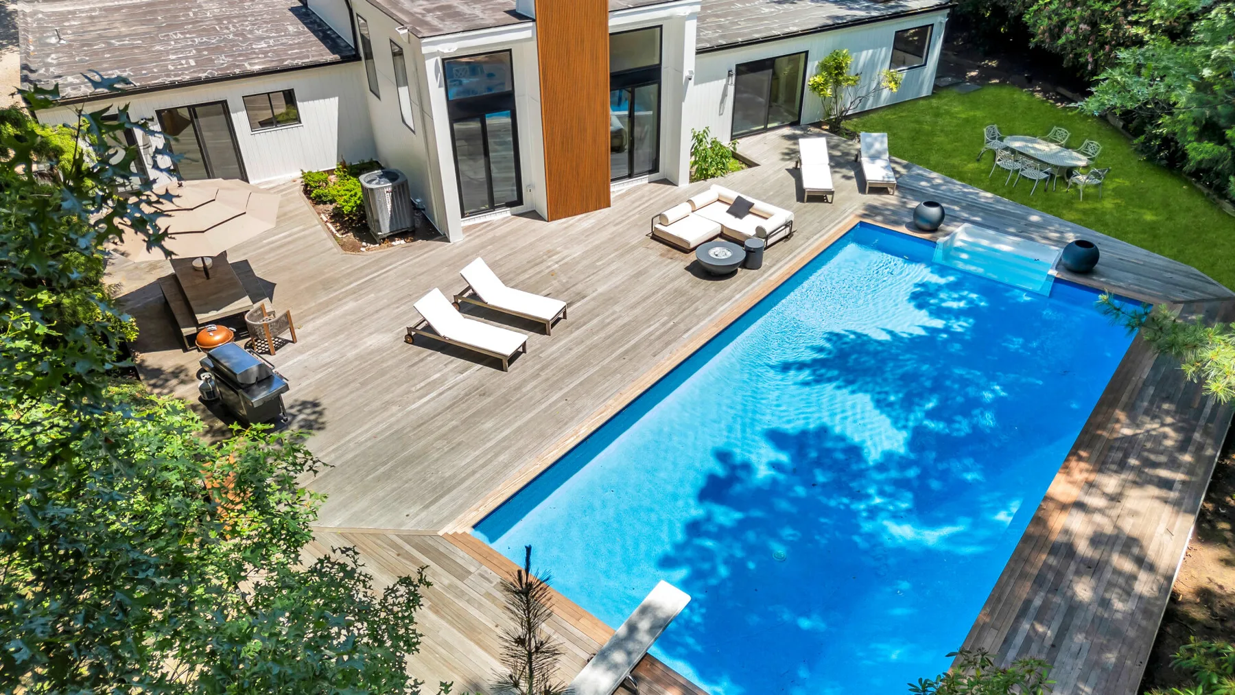 A bright outdoor swimming pool is surrounded by a wooden deck and lounge seating.