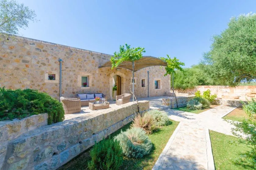 Una casa de piedra da a un patio soleado con asientos al aire libre y un sendero de jardín.