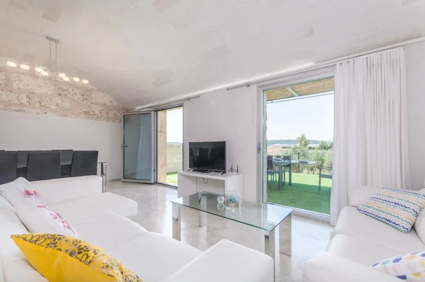 Una sala luminosa con asientos blancos, un televisor y grandes puertas de vidrio.