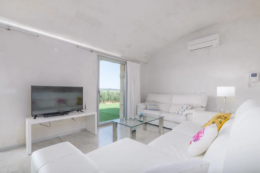 Una sala de estar luminosa con asientos blancos, una mesa de centro de vidrio y un televisor.