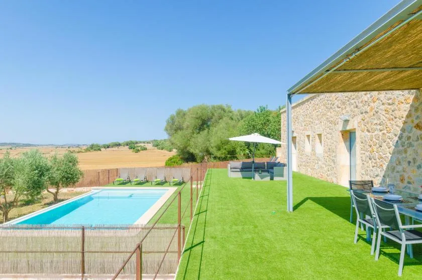 Un patio soleado junto a una piscina, con una mesa y asientos bajo un toldo.