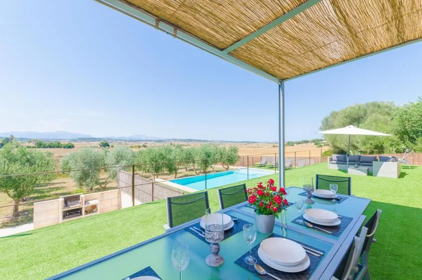 Una mesa de comedor al aire libre está bajo un toldo tejido, con vistas a una piscina y al campo.