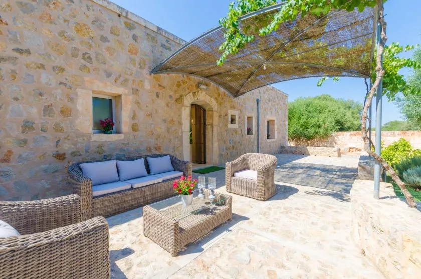 Un patio de piedra con asientos de mimbre está bajo una cubierta de sombra con hojas.