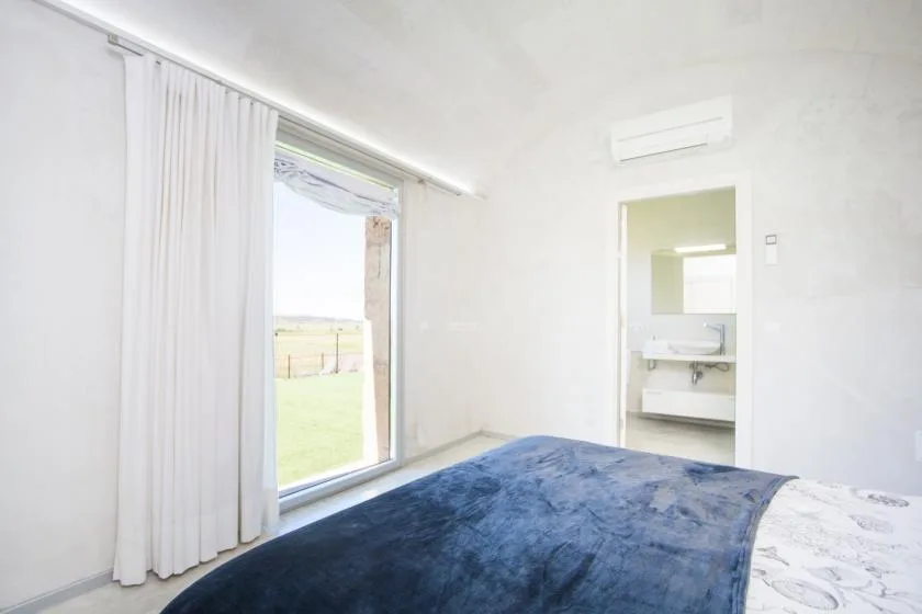 Un dormitorio luminoso con una cama junto a una ventana grande y cortinas blancas.