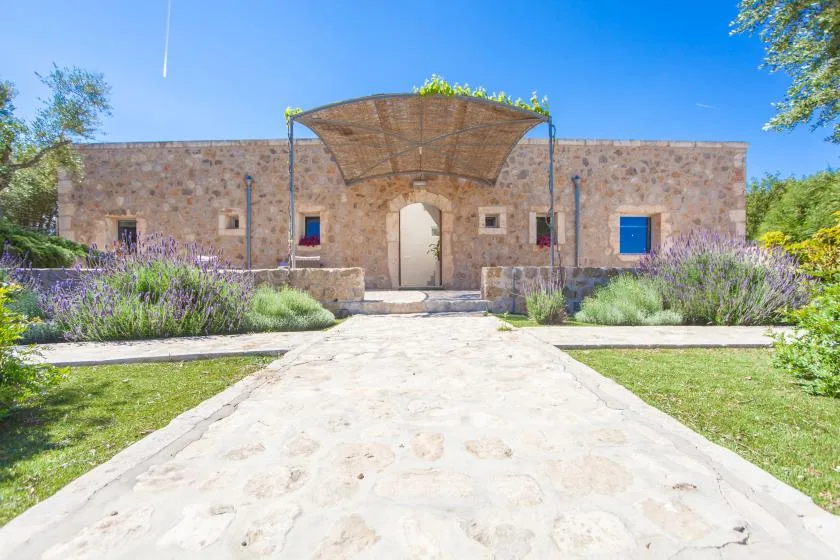Un edificio de piedra con una entrada sombreada se encuentra tras un sendero pavimentado y un jardín.