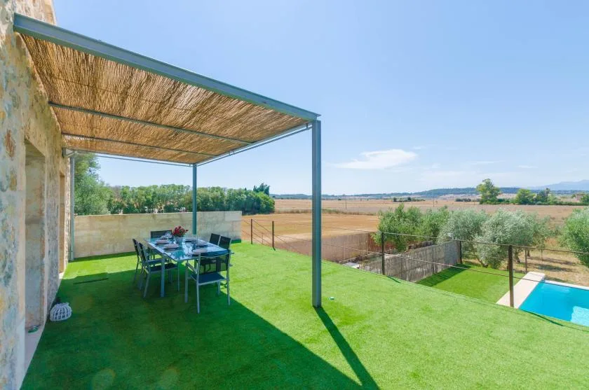 Una zona de comedor exterior con sombra da a un césped verde, campos y una piscina.