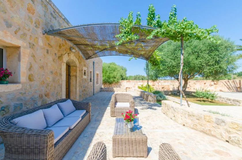 Un edificio de piedra da a un patio soleado con asientos al aire libre y una cubierta frondosa.