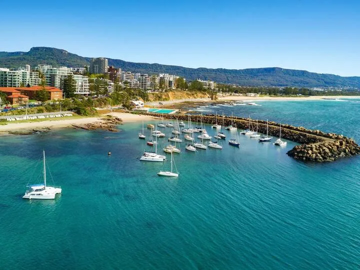 Una bahía costera con veleros anclados junto a un rompeolas rocoso y edificios frente a la playa.