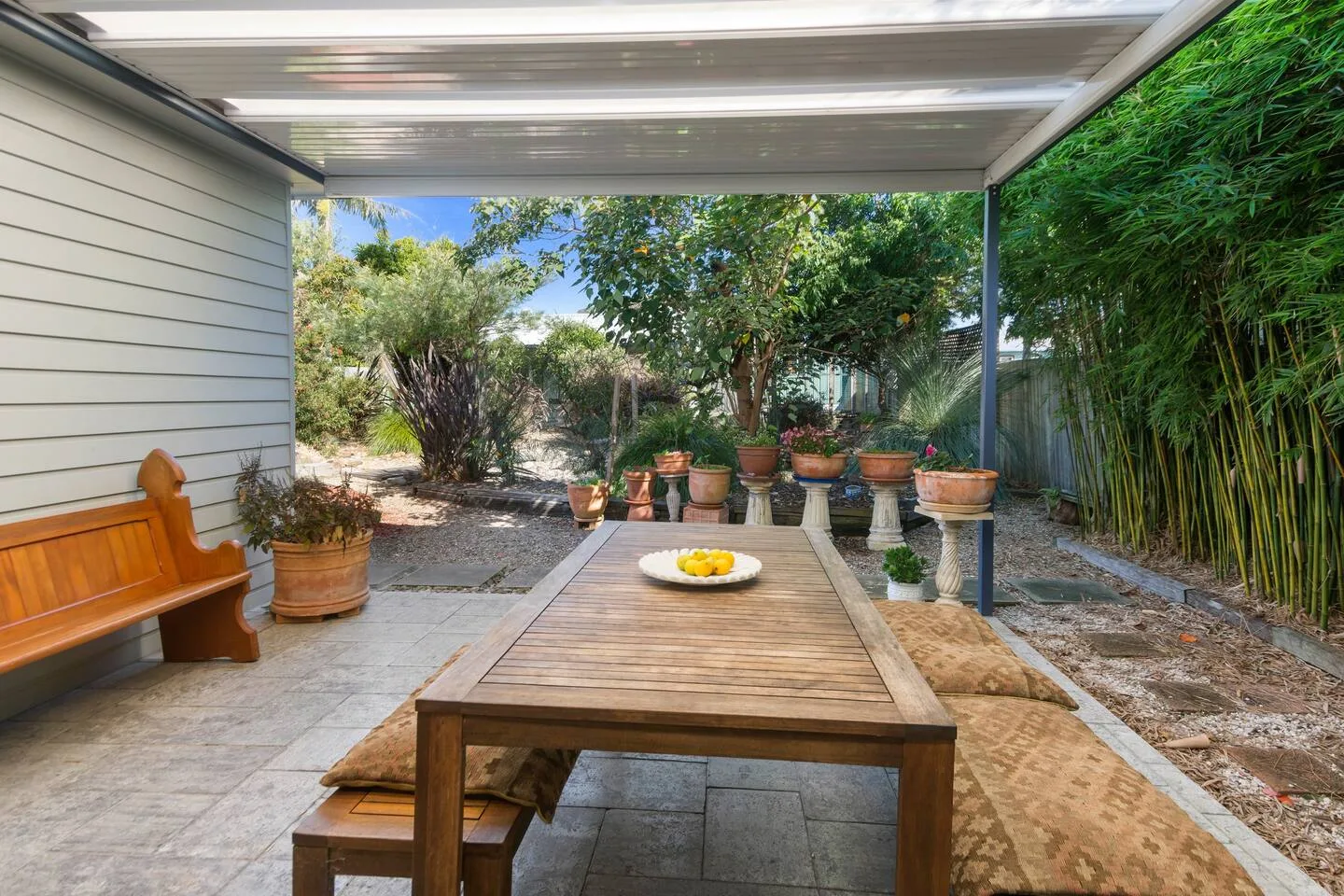 Un patio exterior cubierto tiene una mesa larga de madera, bancos y plantas en macetas.