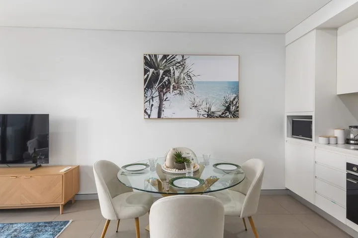 Un comedor luminoso con una mesa de cristal y una lámina enmarcada de la playa.