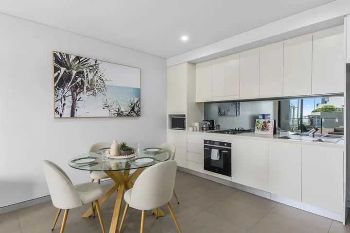 Un comedor luminoso junto a una cocina blanca, con una mesa redonda de vidrio y asientos.