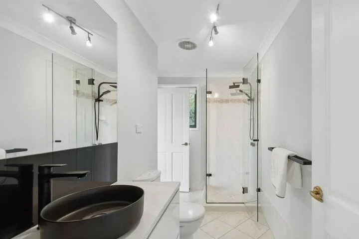 Un baño luminoso con lavabo negro, espejo de pared y ducha de vidrio.