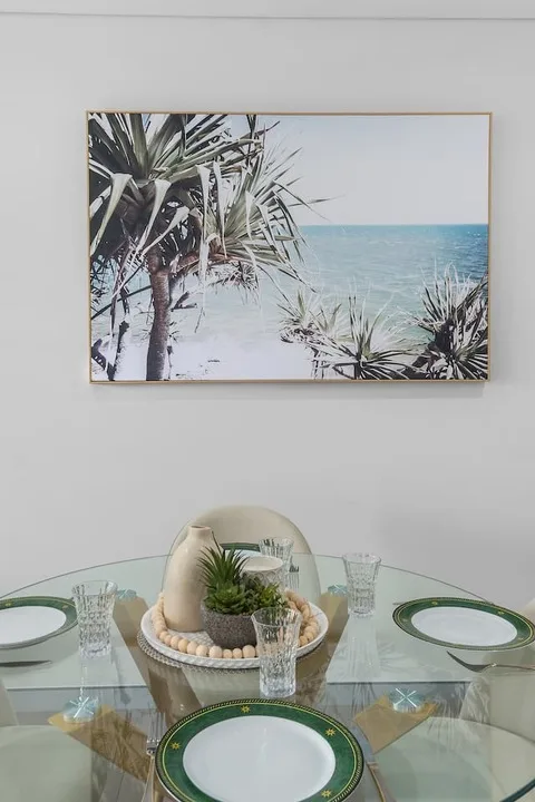 Una mesa de comedor de vidrio está puesta con platos de borde verde bajo una fotografía de playa.