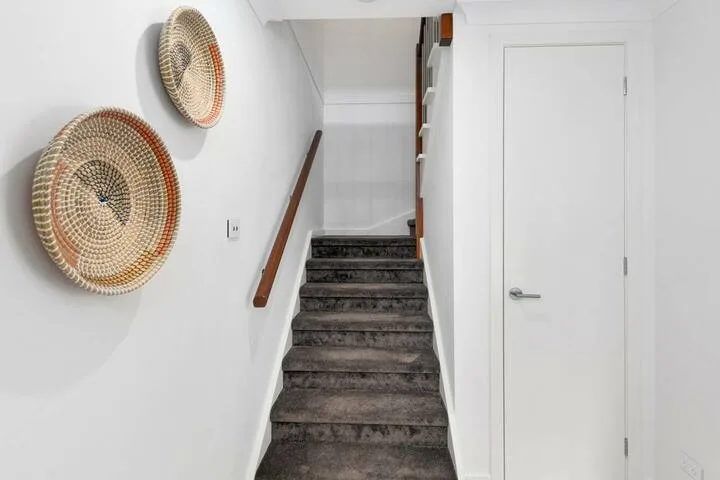 Una escalera interior con peldaños oscuros, pasamanos de madera y adornos tejidos en la pared.