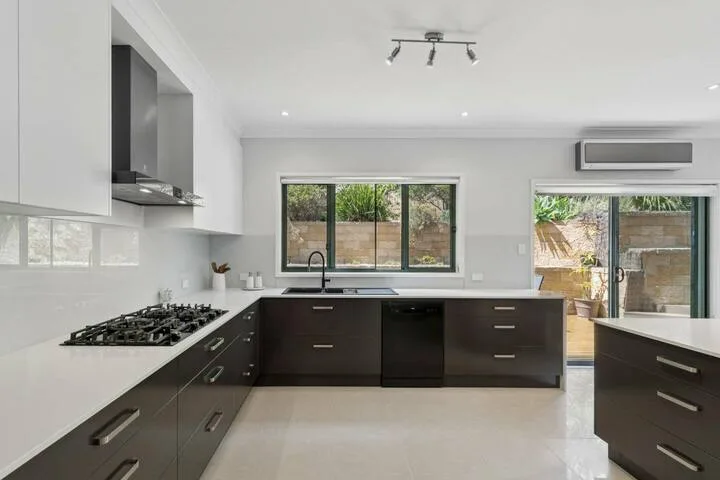 Una cocina moderna con gabinetes oscuros, encimeras blancas y ventanas con vista al jardín.