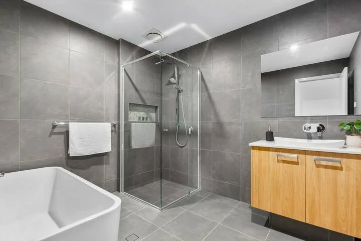 Un baño moderno con paredes de azulejos grises, una ducha de vidrio y un tocador de madera.