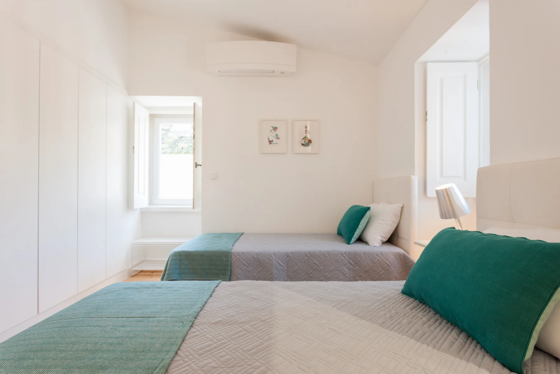 A bright bedroom with two beds, teal bedding, and framed wall art.