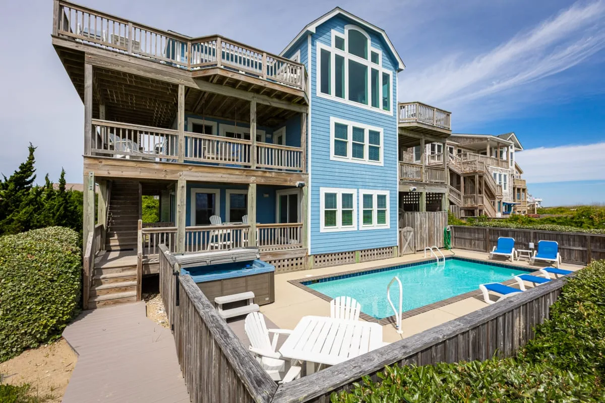 A blue multi-story coastal house with a fenced swimming pool and outdoor seating.