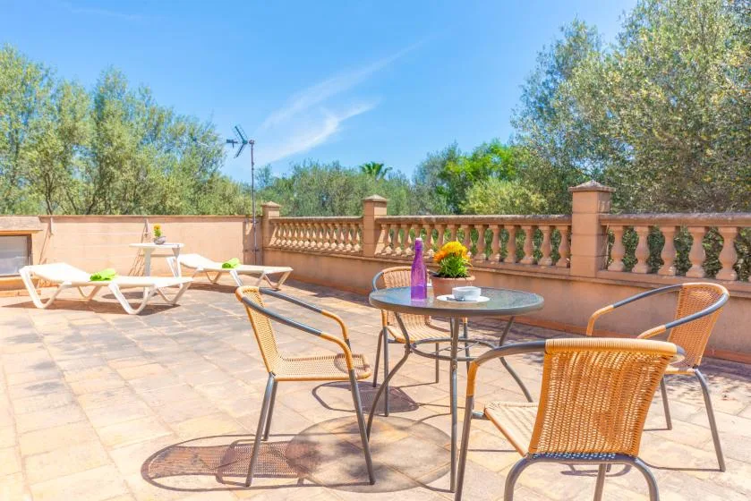 Una terraza soleada tiene una mesa redonda y asientos junto a una barandilla.