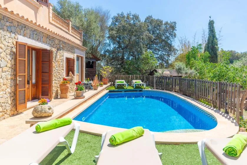 Una piscina luminosa junto a una casa de piedra, con tumbonas y vegetación.