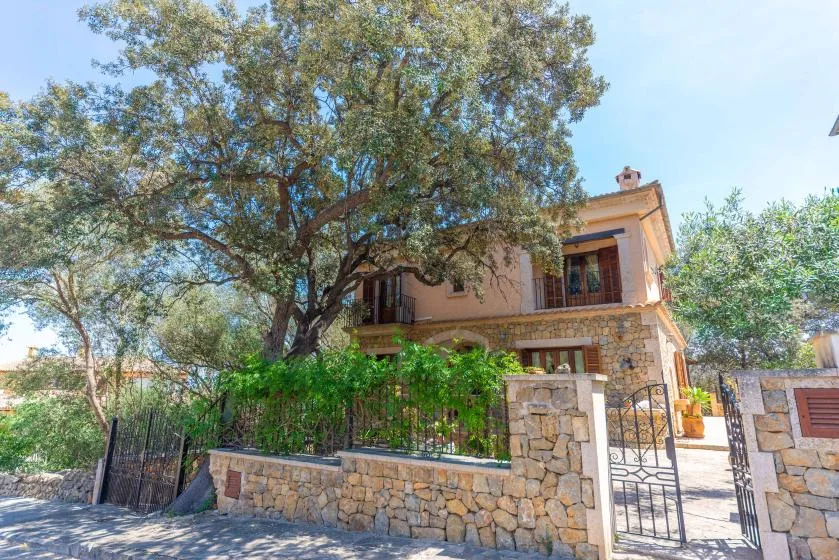 Una casa de dos plantas se alza detrás de un muro de piedra y árboles frondosos.