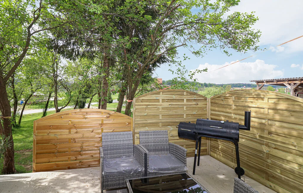 An outdoor patio with seating, wooden fencing, and a barbecue beneath leafy trees.