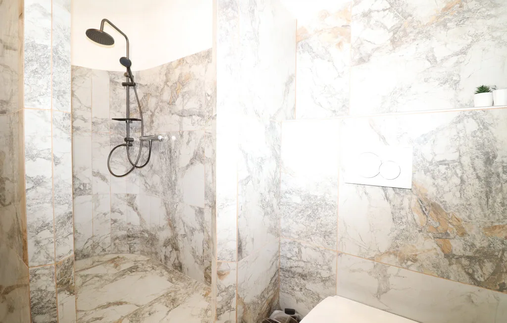 Un baño revestido de mármol con ducha y placa de descarga en la pared.