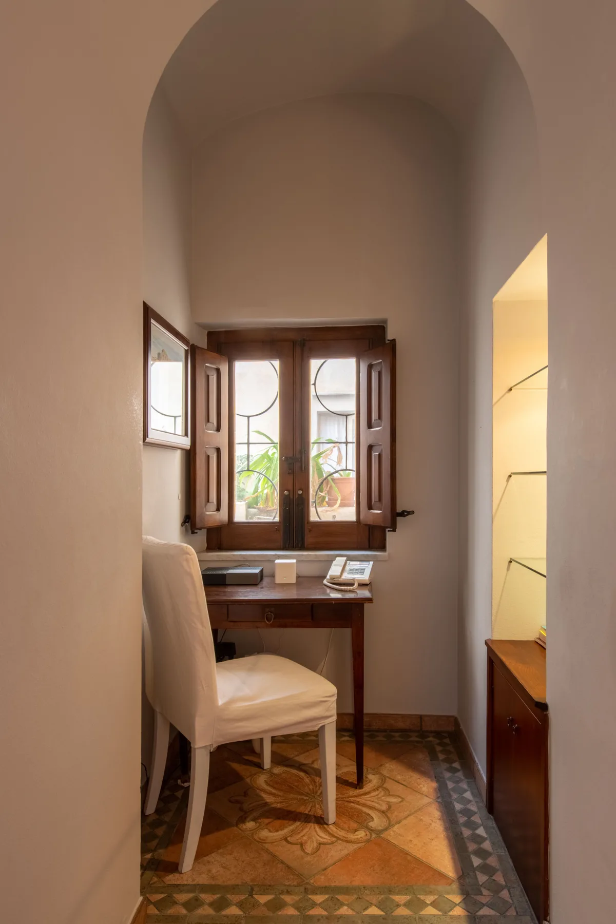 Una habitación pequeña con un escritorio junto a una ventana de madera y suelo de baldosas estampadas.