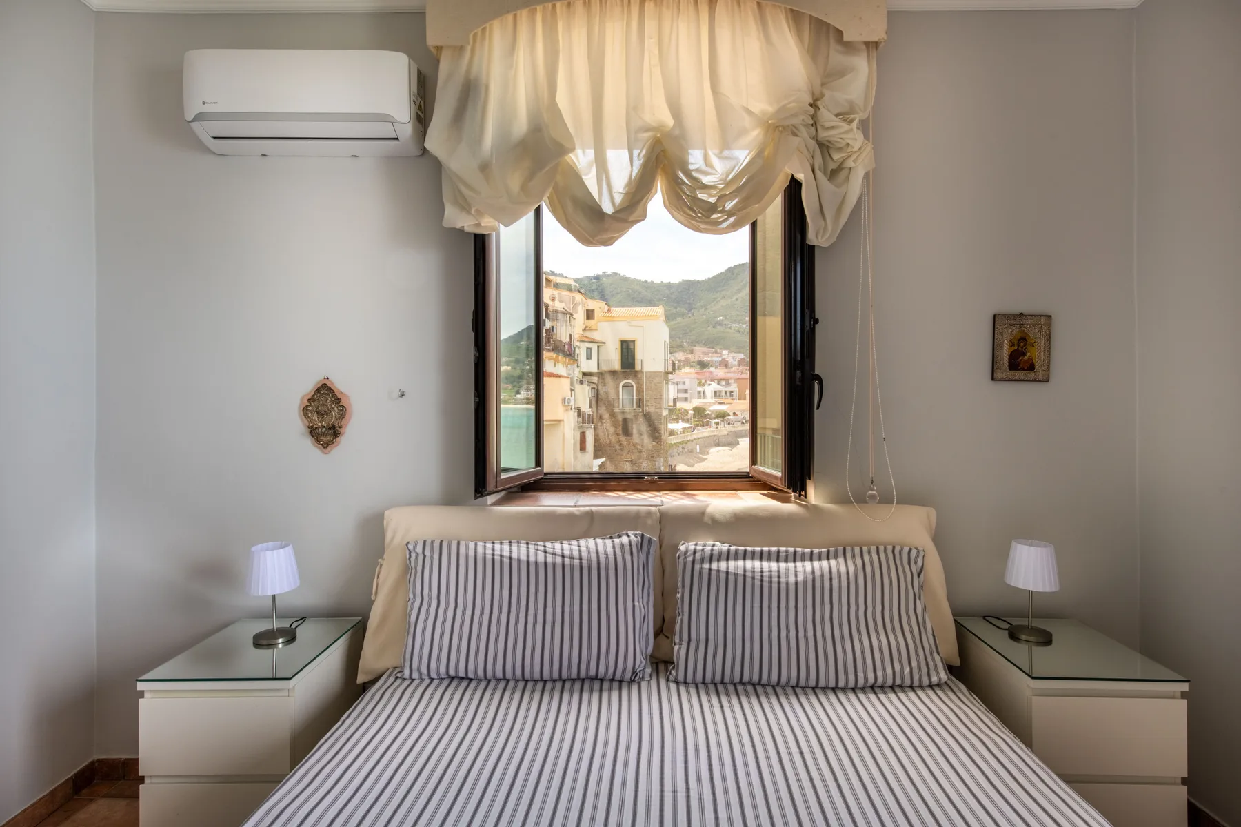 Un dormitorio con una cama bajo una ventana abierta con vistas a edificios y colinas.