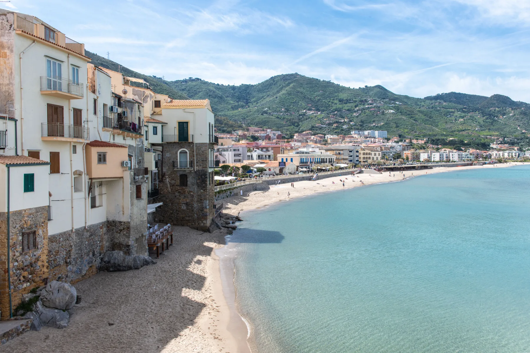 Una playa curva de arena bordea edificios costeros, con colinas verdes detrás del pueblo.