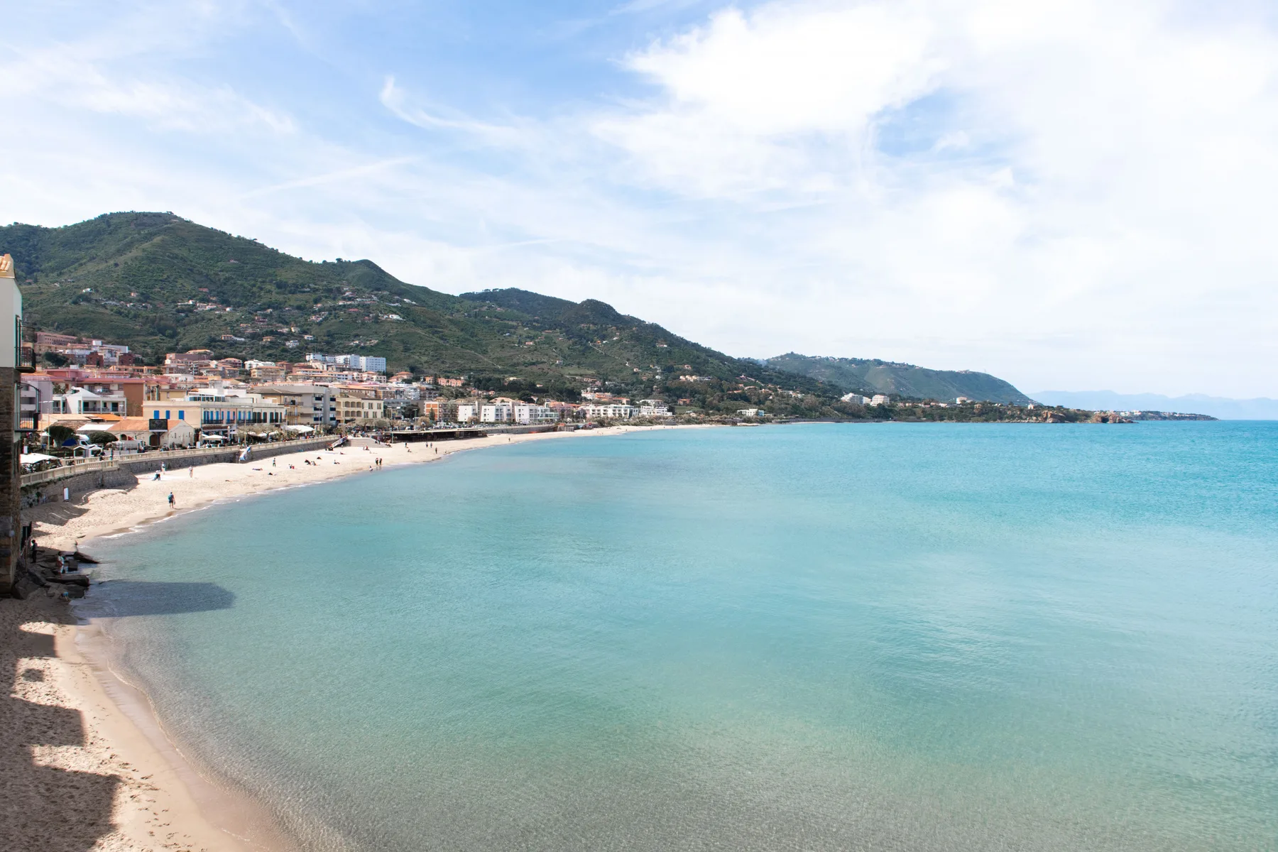 Una playa costera bordea un pueblo, junto a aguas turquesas tranquilas y colinas verdes.