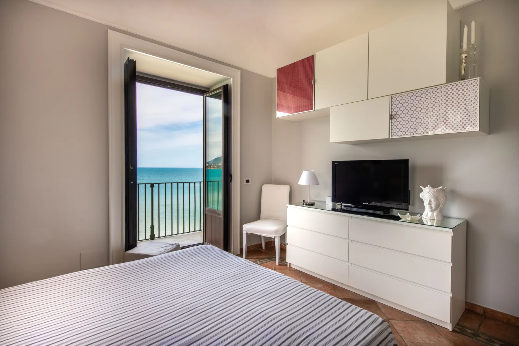Un dormitorio con una cama, un televisor y una puerta abierta al balcón con vistas al mar.