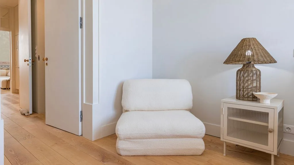 Una zona de estar luminosa con un asiento bajo tapizado junto a un pequeño mueble y una lámpara.