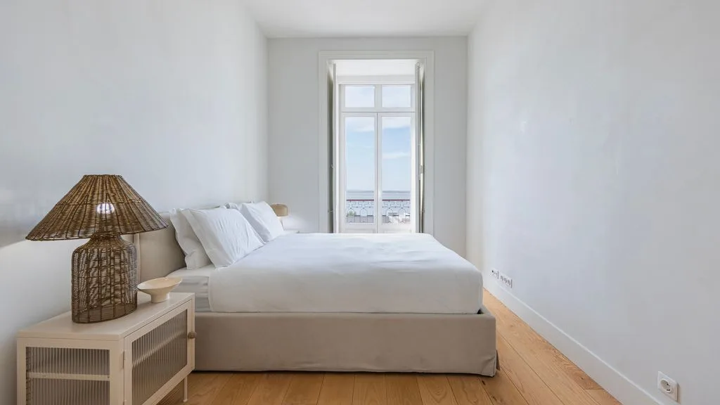 Un dormitorio luminoso con una cama, una lámpara tejida y una ventana alta.