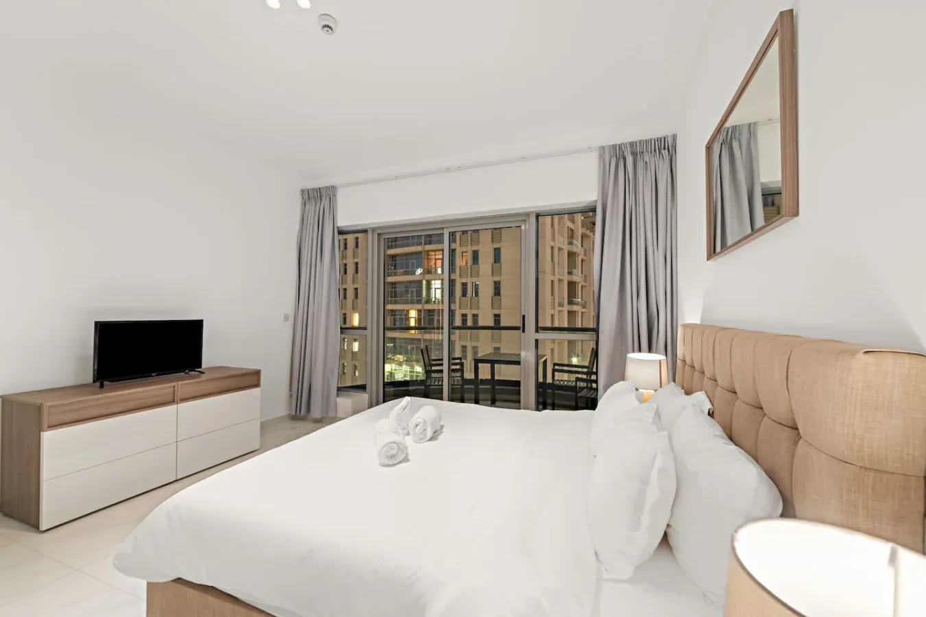 Un dormitorio luminoso con una cama bien hecha, un televisor y grandes ventanas con cortinas.