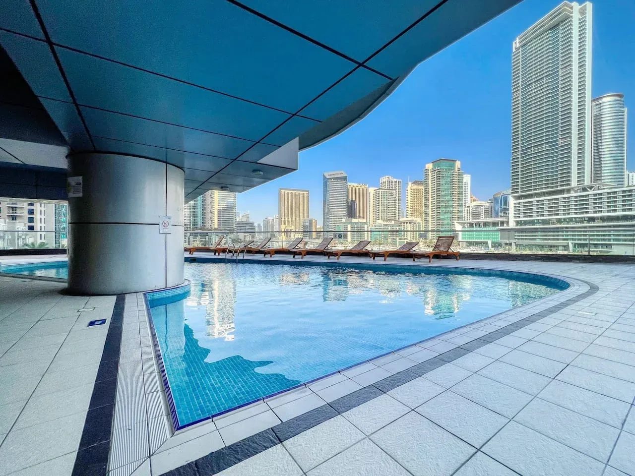 Una piscina en la azotea está bajo un toldo azul, con edificios altos al fondo.