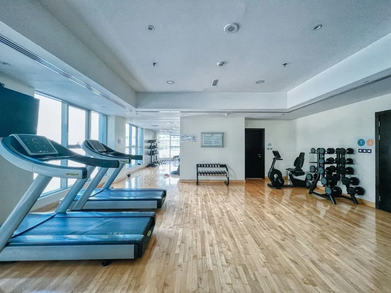 Un gimnasio con cintas de correr junto a las ventanas y equipos de ejercicio junto a la pared.