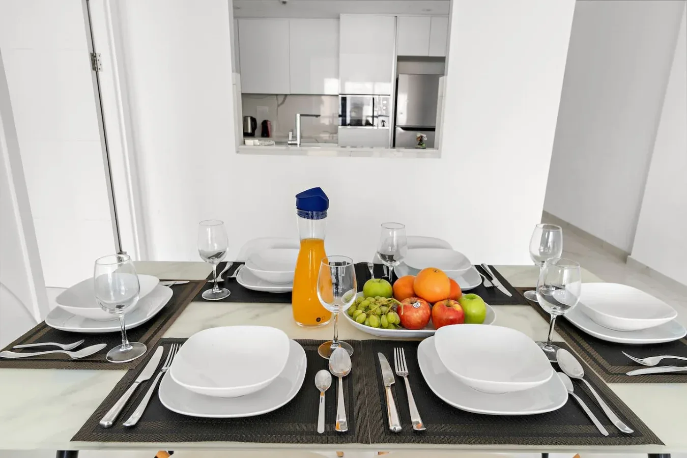 Una mesa de comedor está preparada con platos, vasos, cubiertos y un frutero.
