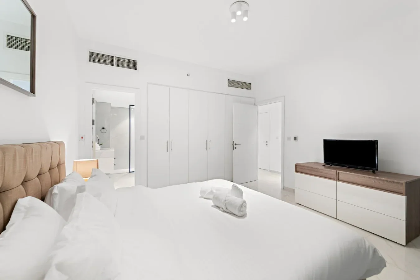 Un dormitorio luminoso con ropa de cama blanca, un cabecero acolchado y un televisor.