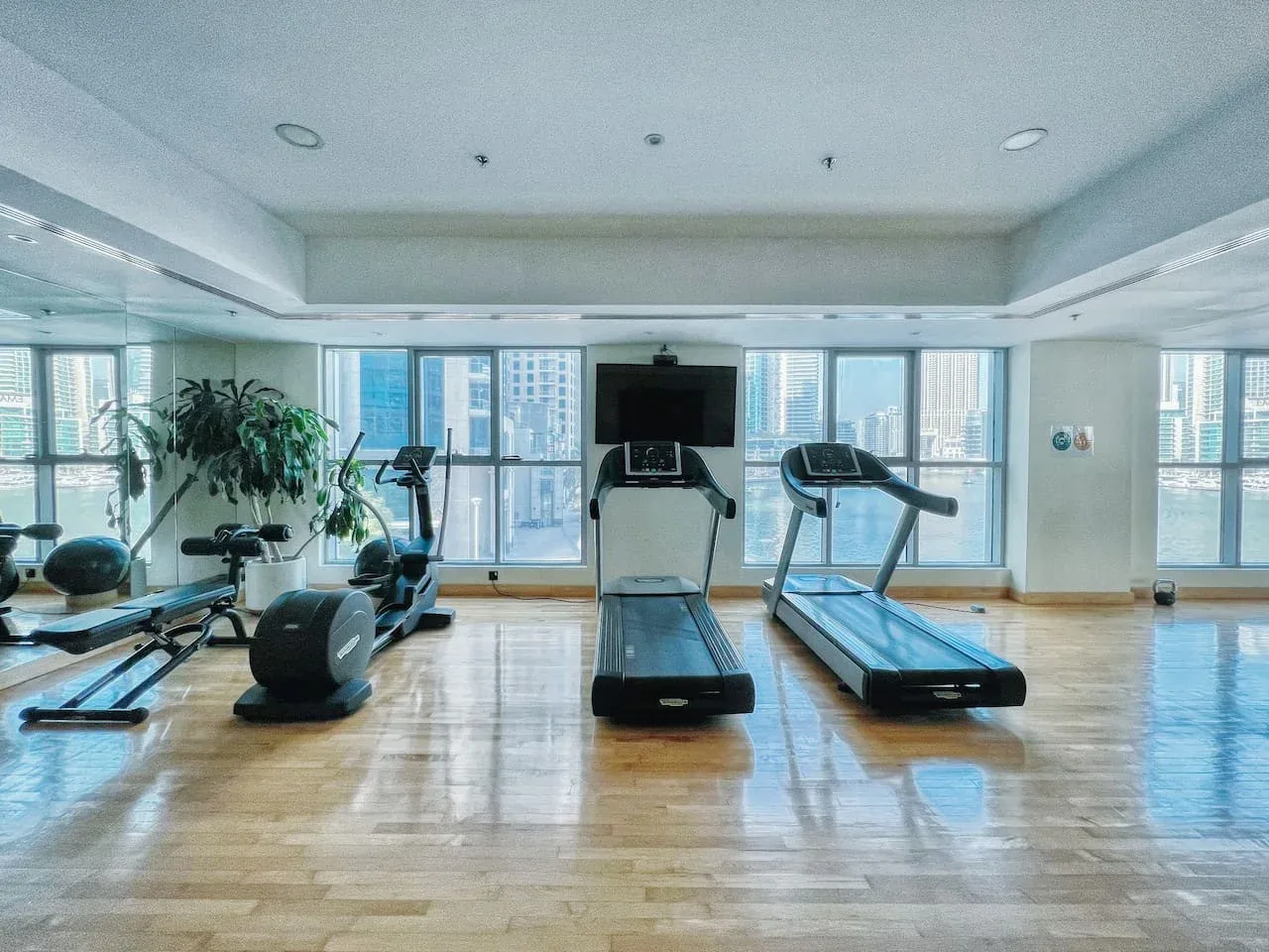 Un gimnasio luminoso con máquinas de ejercicio y grandes ventanales con vistas a edificios de la ciudad.