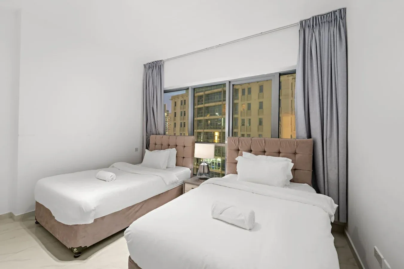 Un dormitorio con dos camas, cortinas grises y una gran ventana con edificios a la vista.