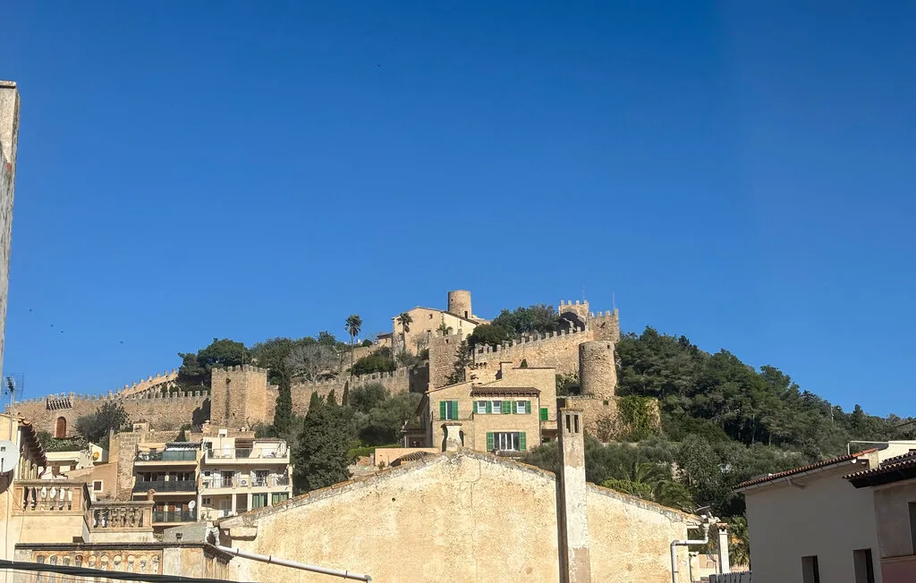 Un castillo en una colina se alza sobre edificios agrupados bajo un cielo azul despejado.